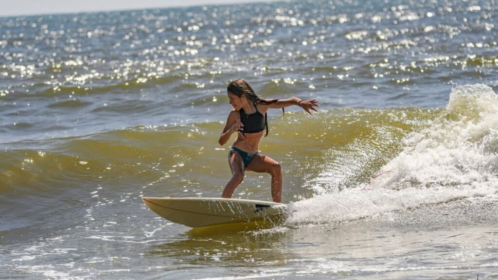 Imagem mostra uma criança de dez anos, com diabetes tipo 1, branca, loira com um macacão próprio para entrar no mar. Na imagem ao fundo tem um mar com ondas e a criança surfando em uma prancha.