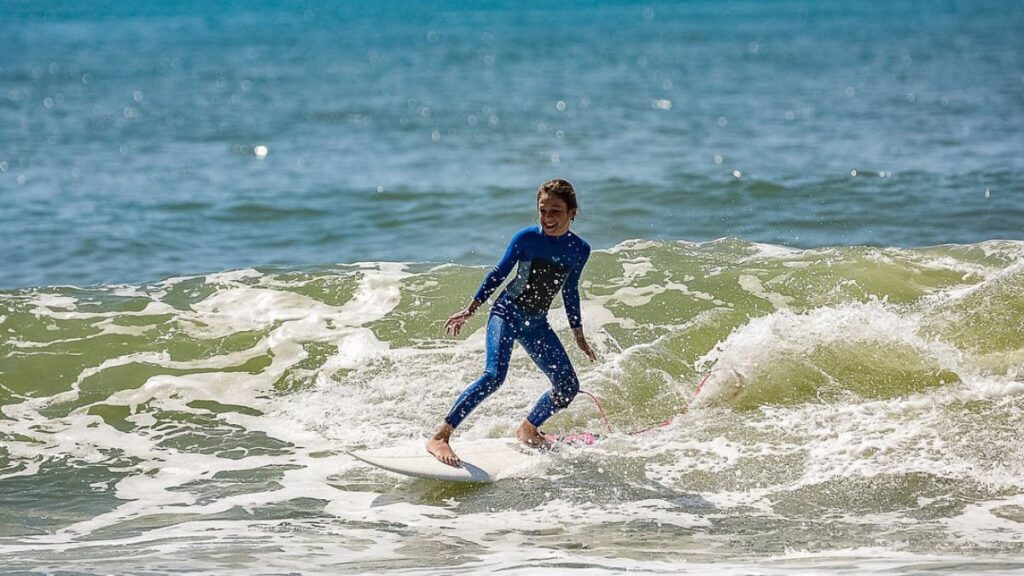 Imagem mostra uma criança de dez anos, com diabetes tipo 1, branca, loira com um macacão próprio para entrar no mar. Na imagem ao fundo tem um mar com ondas e a criança surfando em uma prancha.