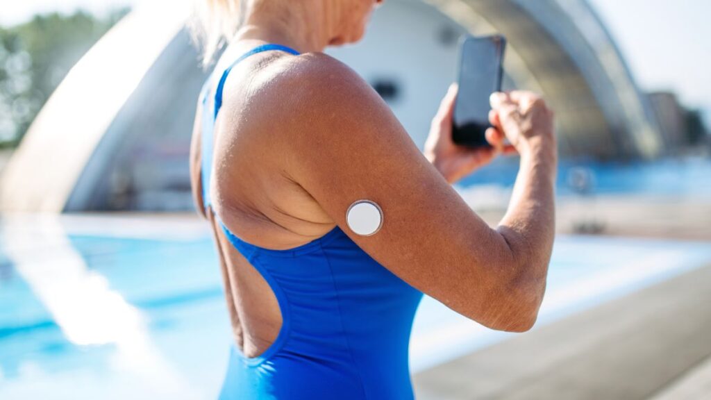 Quem tem diabetes pode nadar? Especialista revela como a glicose fica após a natação 1 Mulher com sensor de glicose no braço usando celular à beira da piscina antes de fazer natação com diabetes