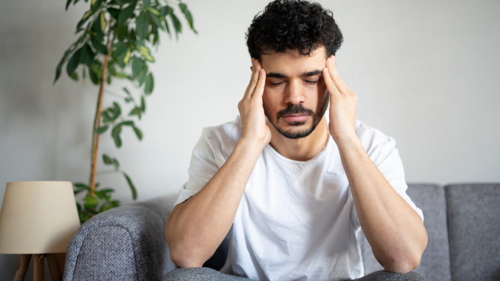 Diabetes pode causar dor de cabeça? Entenda quando a glicose pode estar por trás do sintoma Homem sentado no sofá com dor de cabeça e as mãos nas têmporas