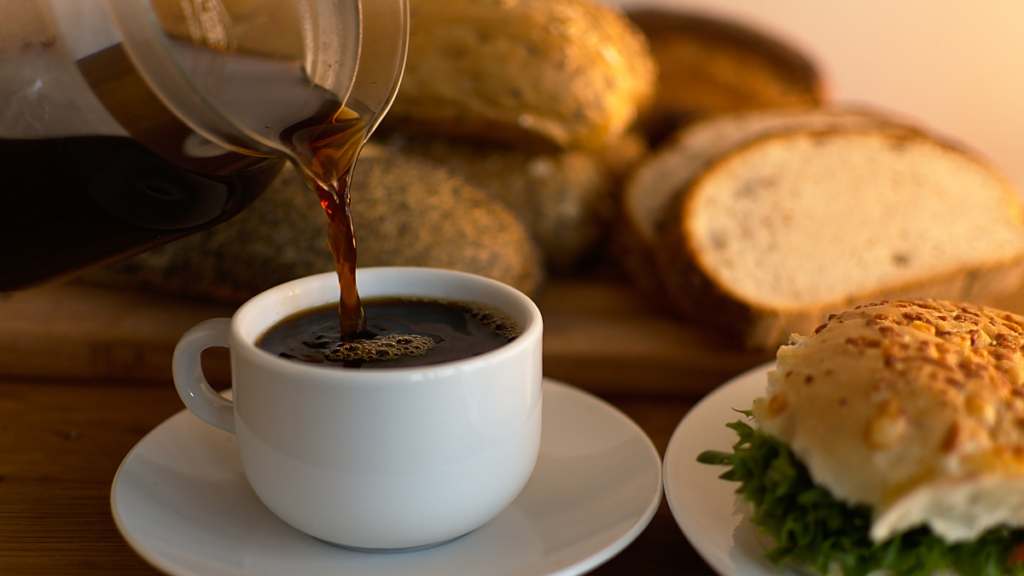 Café da manhã e diabetes: o horário da primeira refeição faz diferença? Café sendo servido em uma xícara branca com pães e sanduíche, em contexto de alimentação para diabetes.