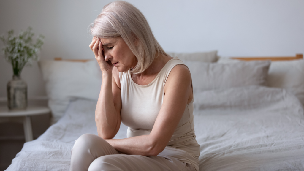 Menopausa e diabetes: o que acontece com o controle da glicose nessa fase? Mulher na menopausa sentada na cama com a mão na testa, ilustrando os desafios dessa fase para quem tem diabetes.